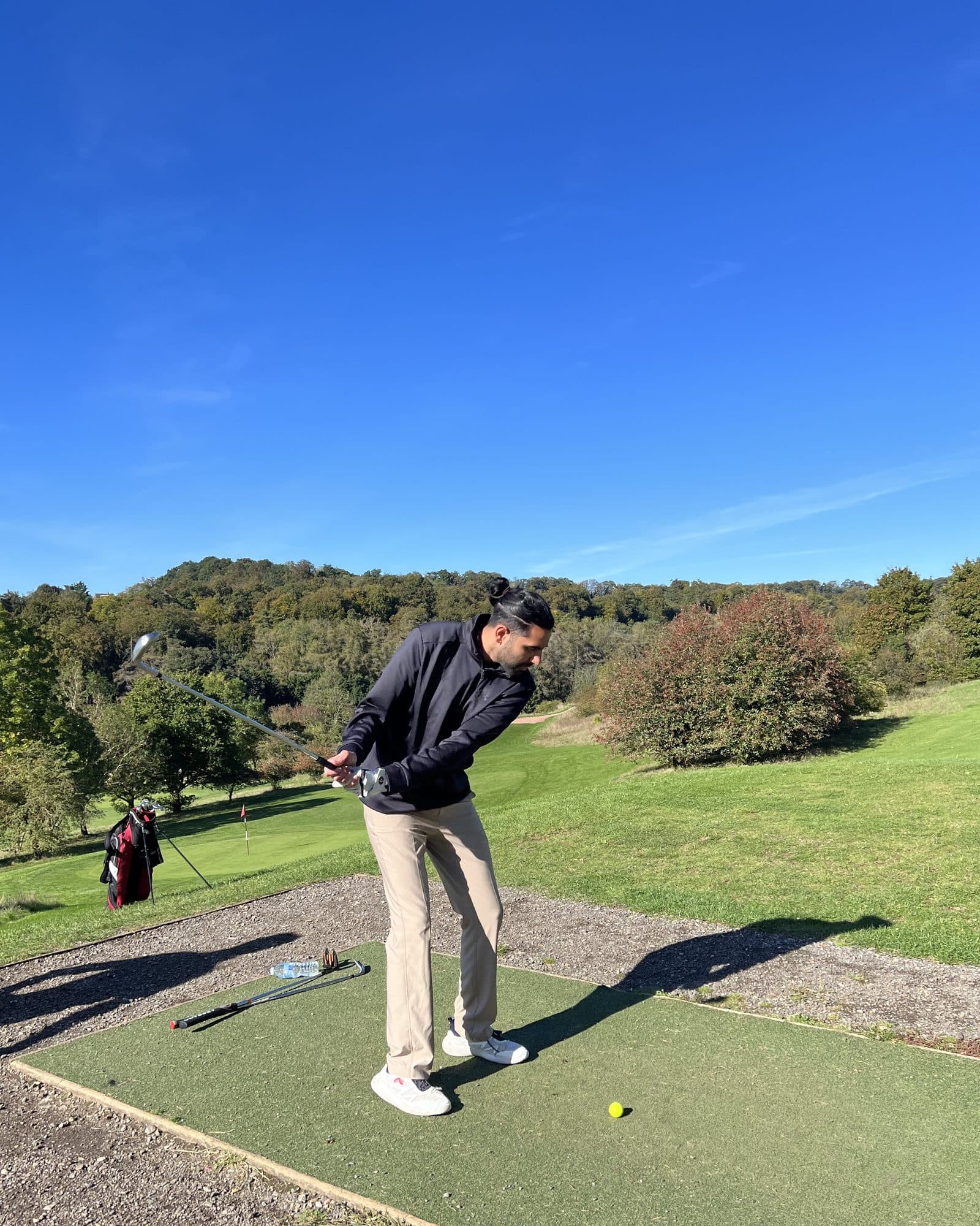 Param in a navy quarter-zip and khaki trousers, hair tied back, halfway into his backswing on a tee mat in bright sun, a wooded hillside and a flag on the green behind him