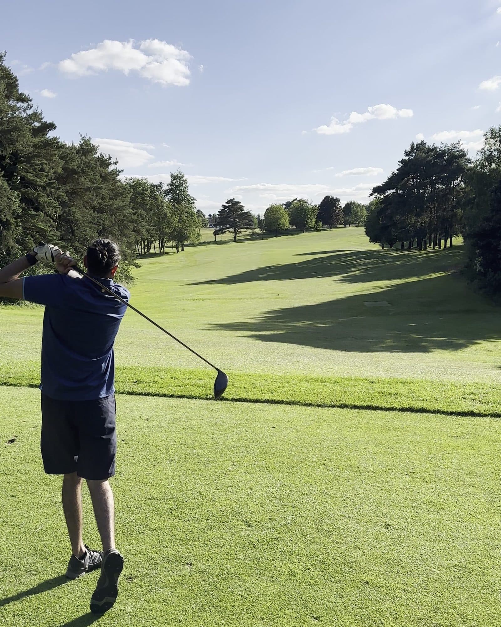 Param from behind in a navy polo and black shorts holding his finish on a tee, a long tree-lined fairway running away under a blue sky