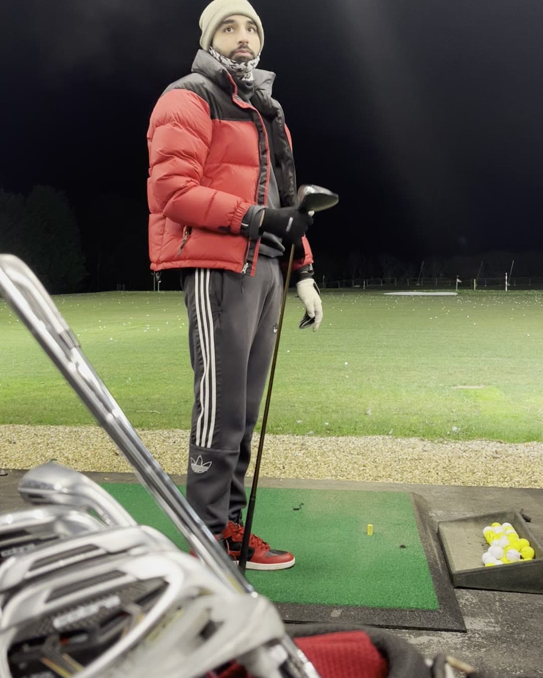 Param standing on the range mat in a red puffer jacket, beanie and scarf, holding a driver and looking off to the side
