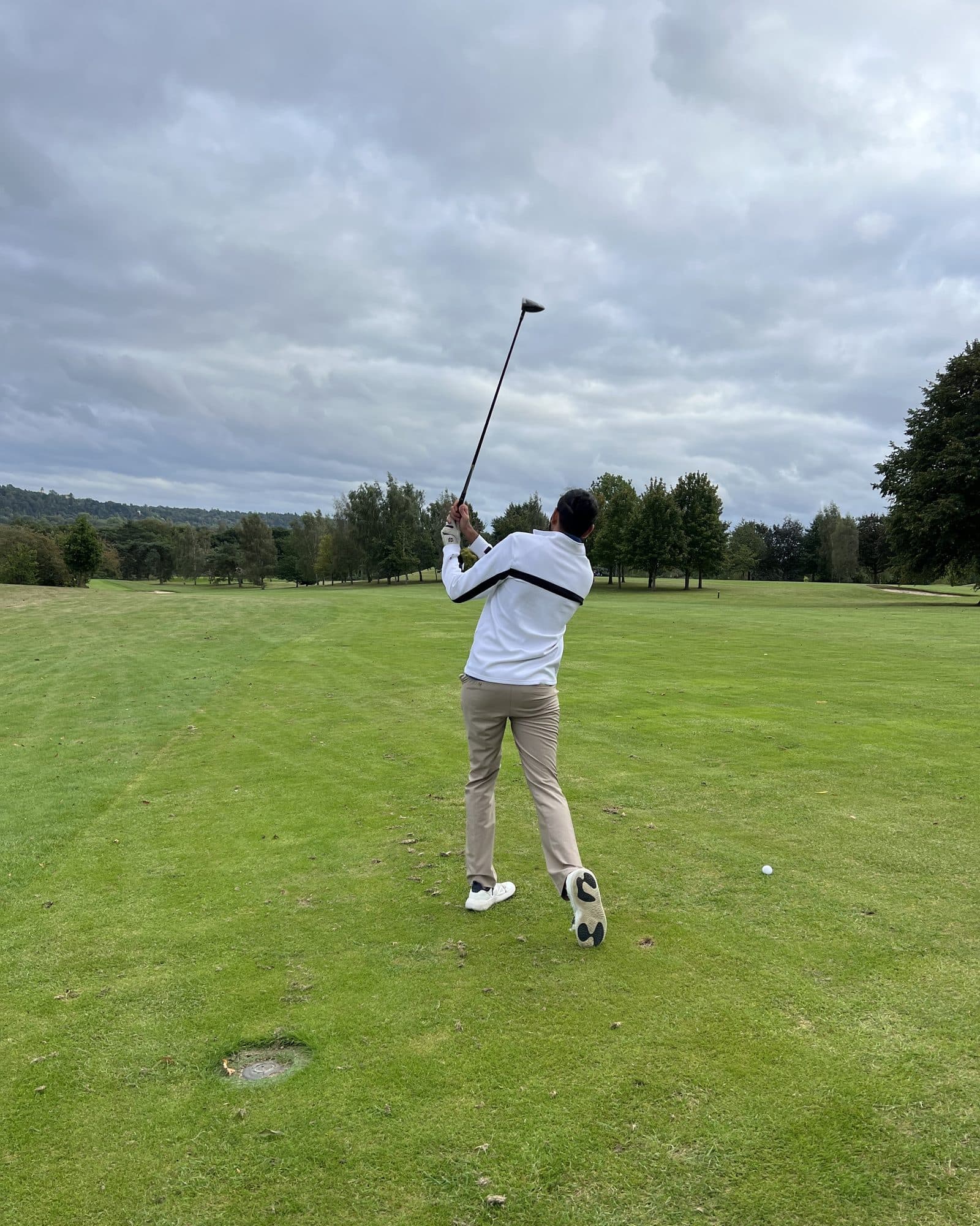 Param from behind in a white jacket and khaki trousers, club high over his shoulder after a swing, the ball still sitting on the fairway beside him under a grey sky