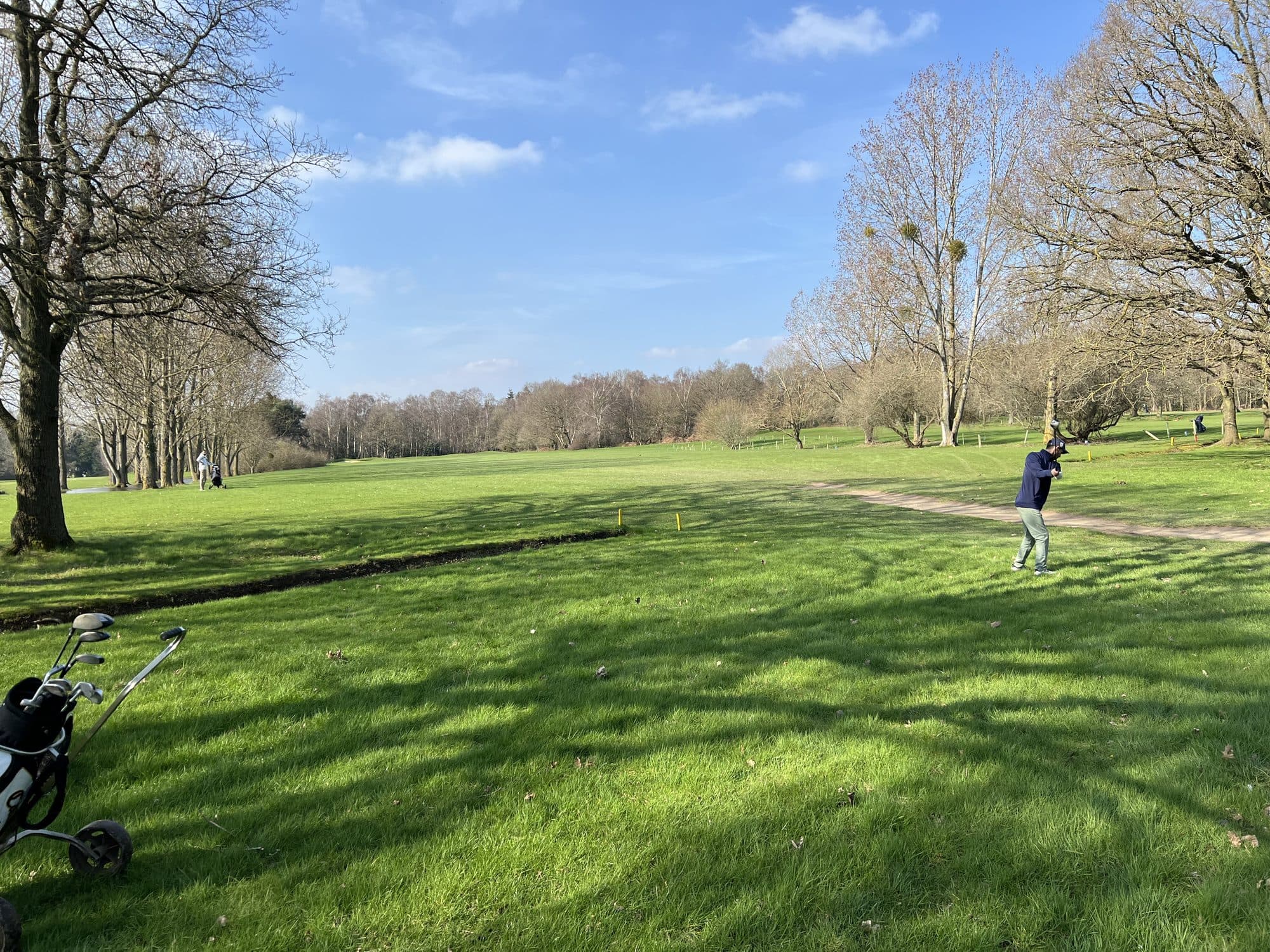 A wide spring fairway in sunshine with bare trees casting long shadows across the grass, a golfer mid-swing on the right and a trolley bag in the left foreground
