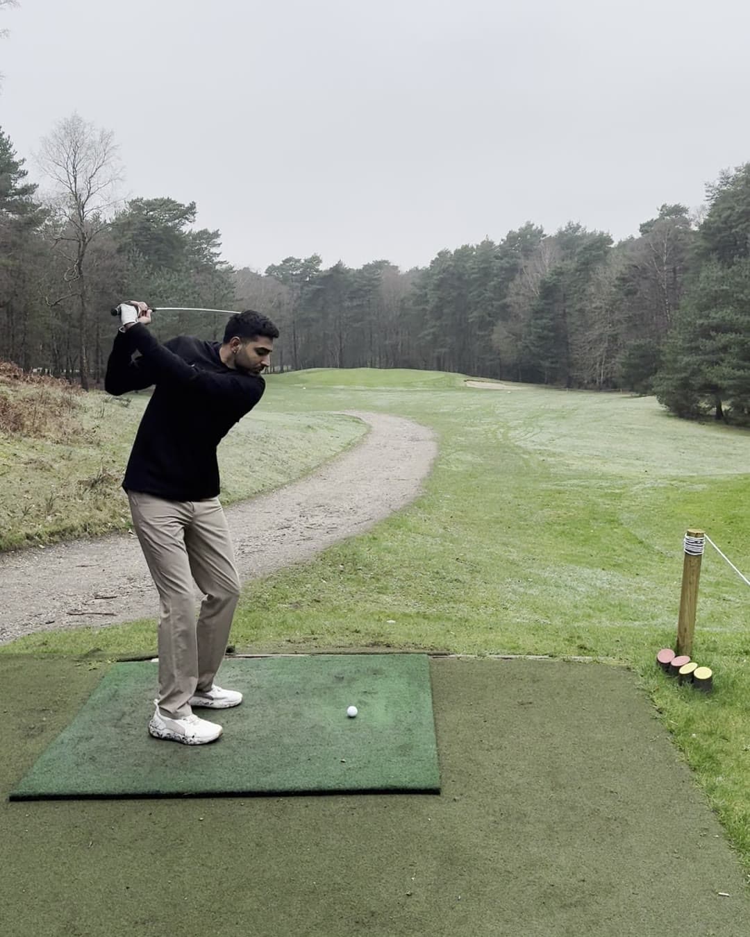 Param in a black hoodie and khaki trousers at the top of his backswing on a tee box, a frosty fairway curving away between pine trees