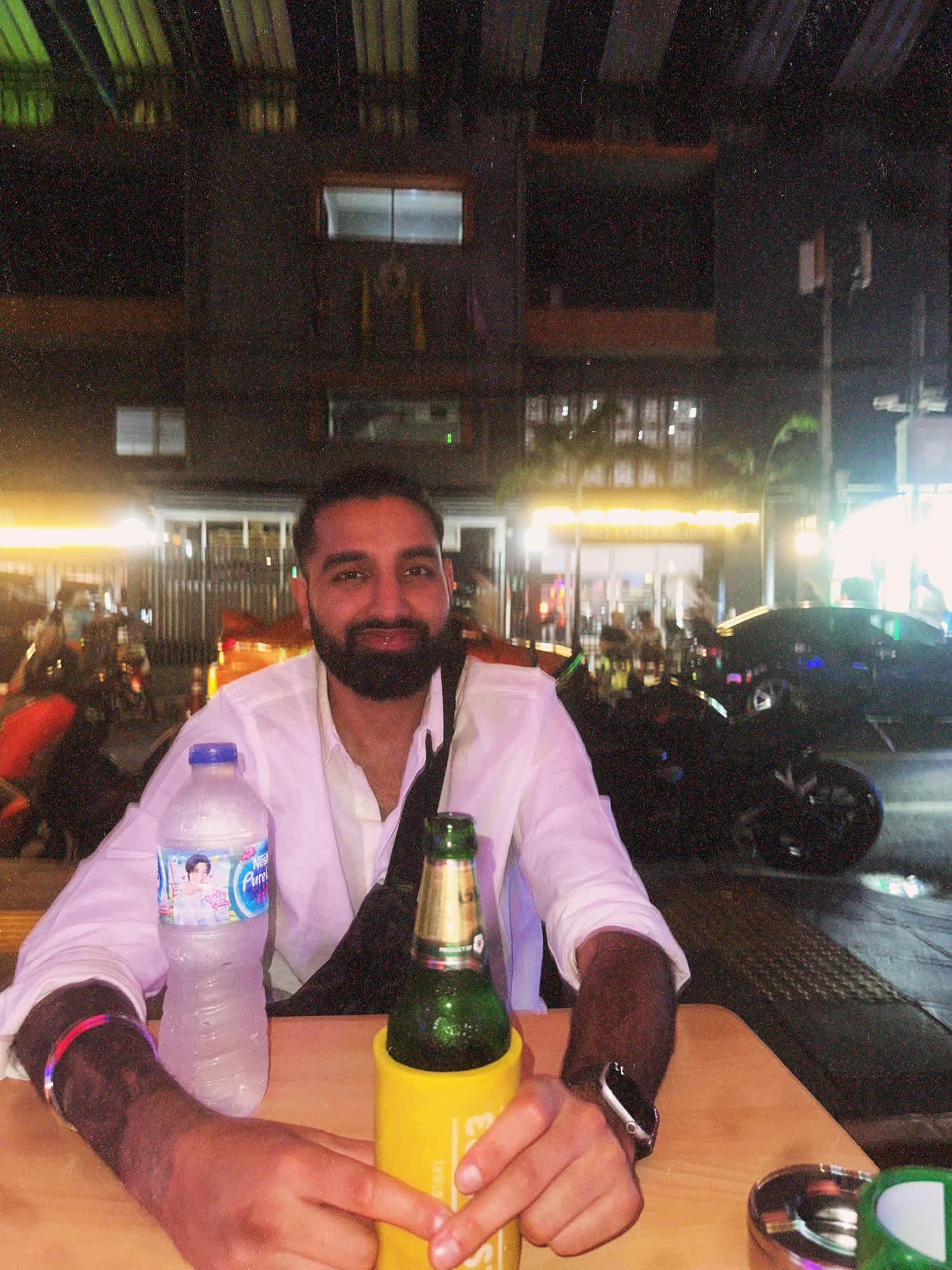 Param in a white shirt smiling at a street-side table at night, a beer in a yellow sleeve in front of him