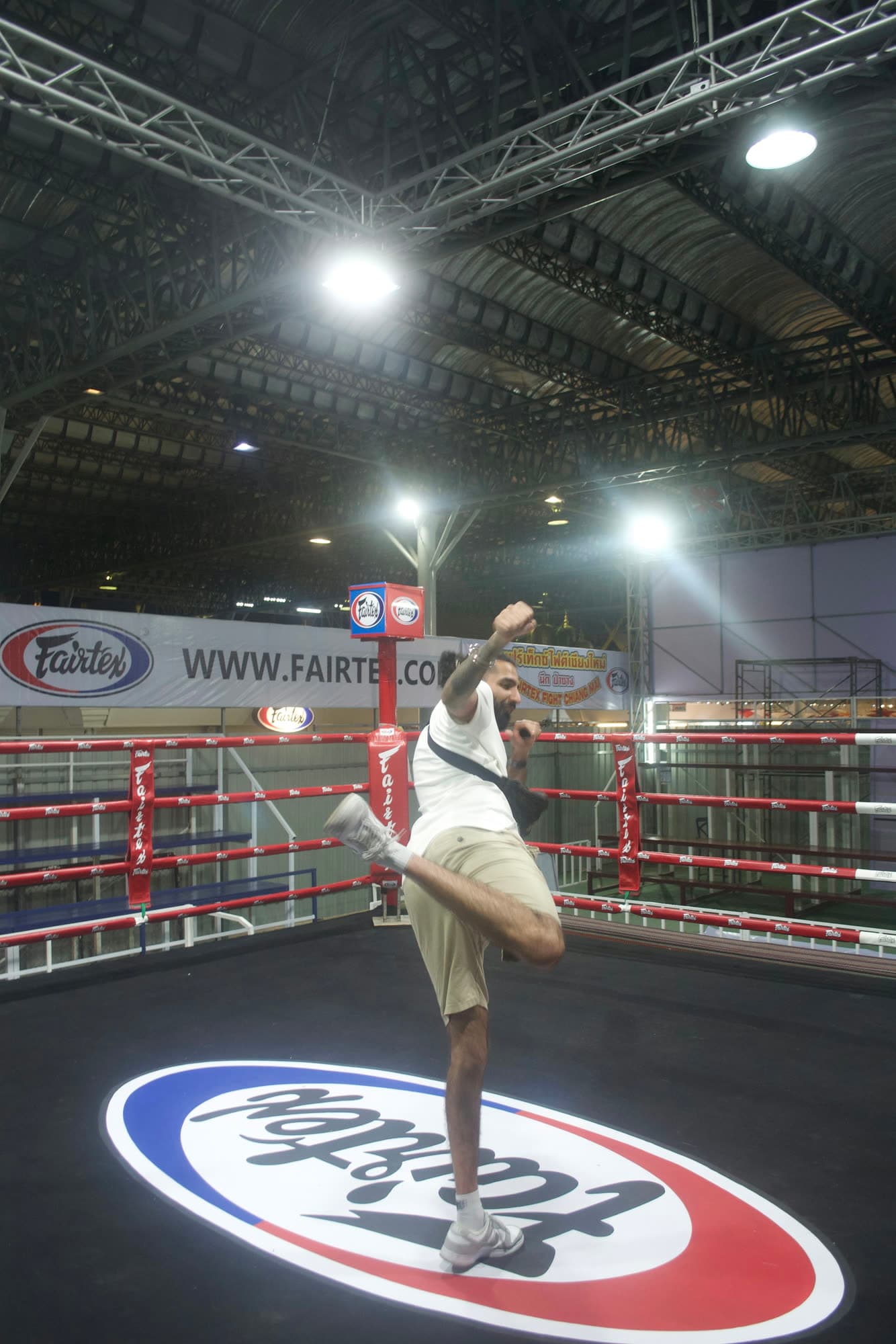 Param in a white T-shirt and shorts throwing a knee in the middle of an empty boxing ring under arena lights