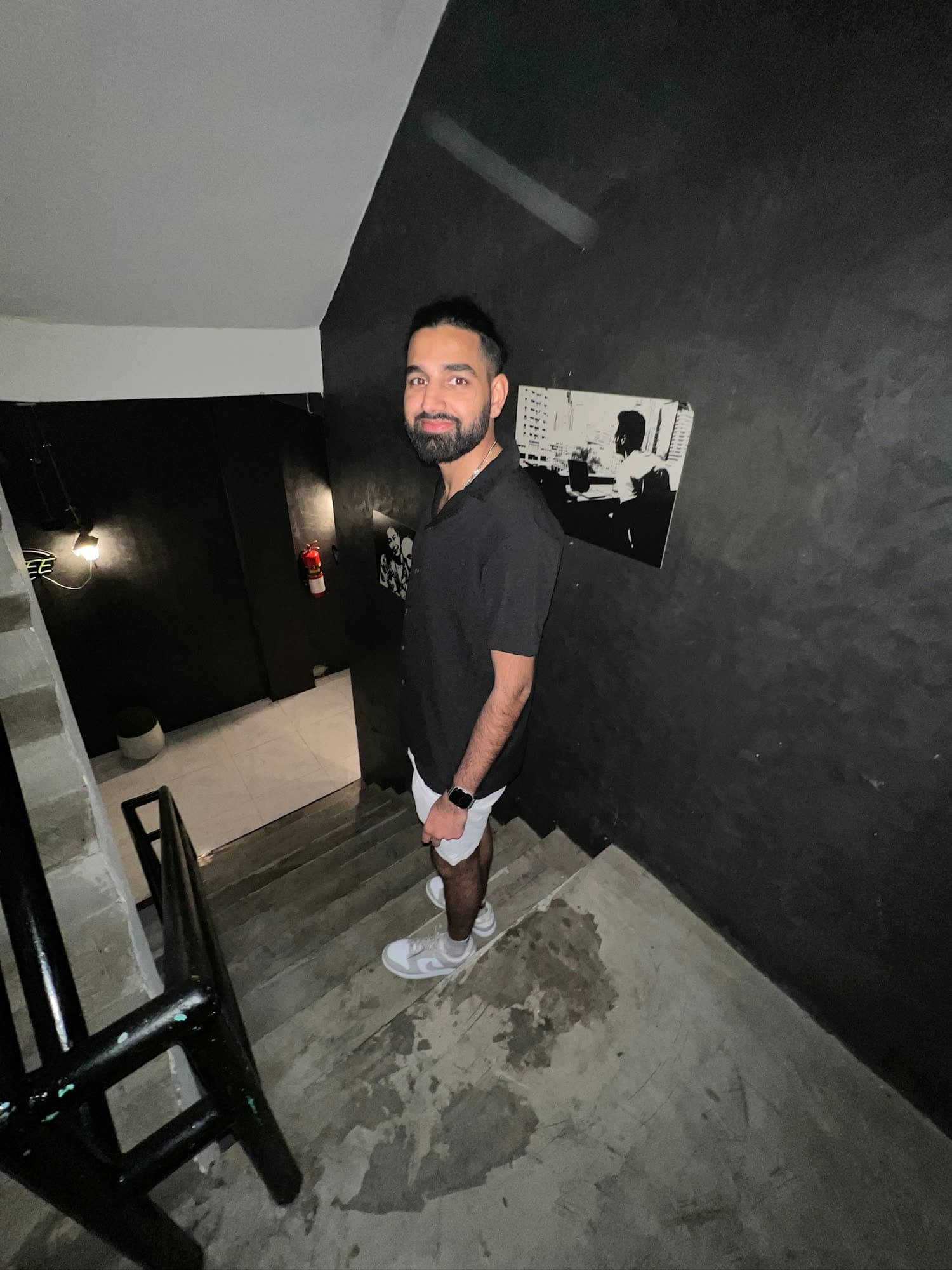 Param in a black shirt and white shorts standing on a concrete stairwell with black walls, looking back over his shoulder