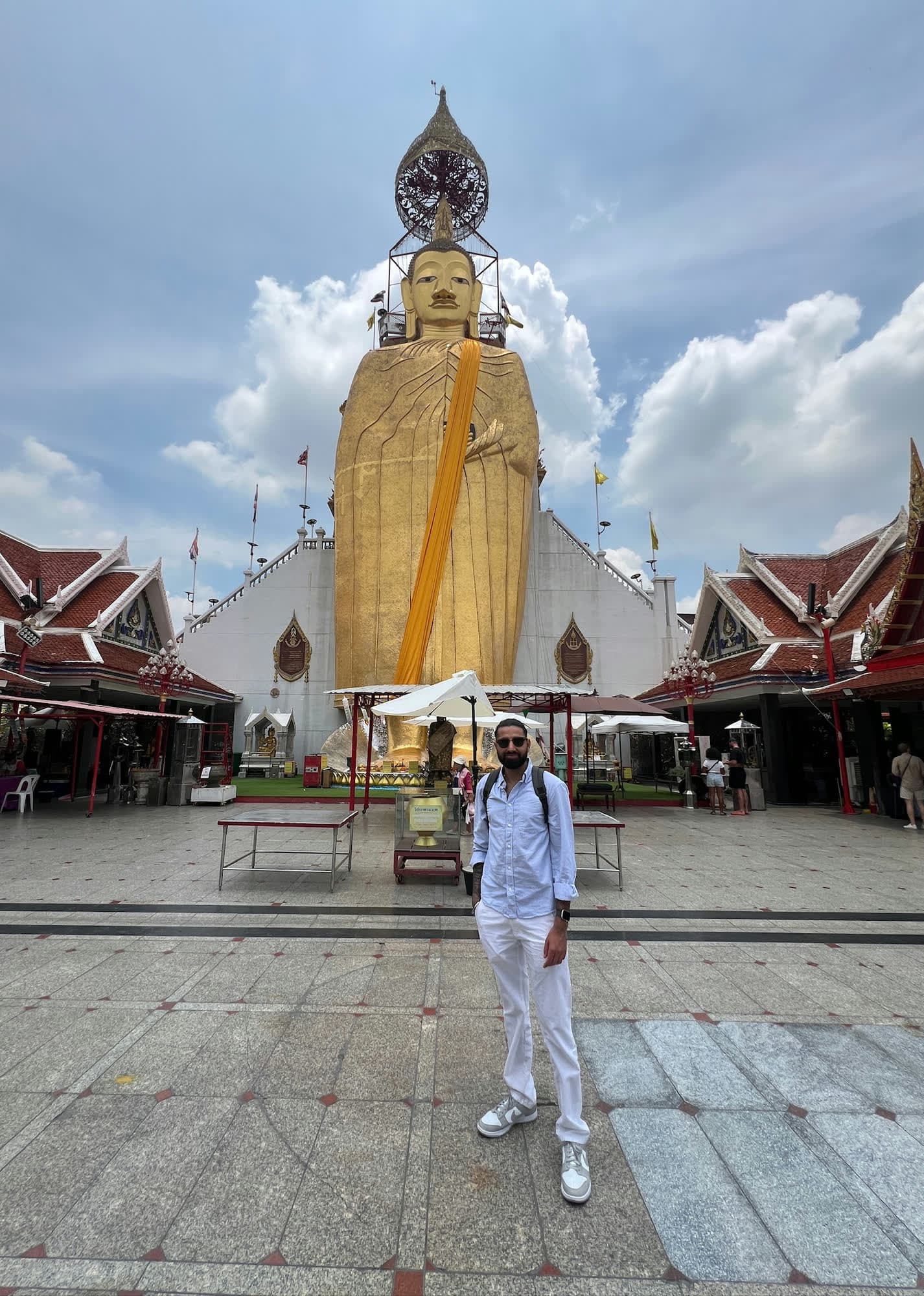 Param in sunglasses, a pale blue shirt and white trousers standing in a temple courtyard below a giant gold standing Buddha