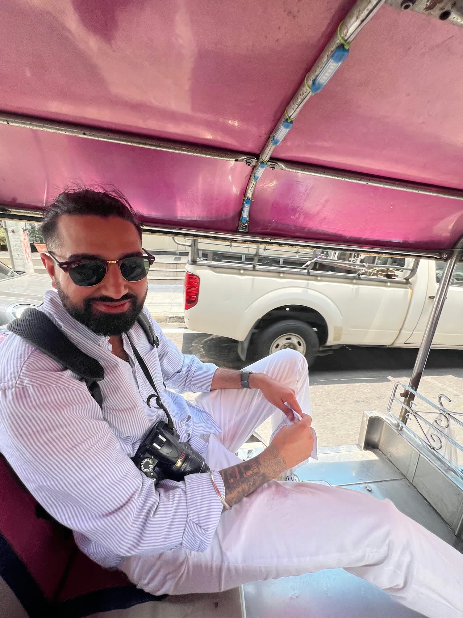 Param in sunglasses and a striped shirt sitting in the back of a pink-roofed tuk-tuk, a camera on his chest