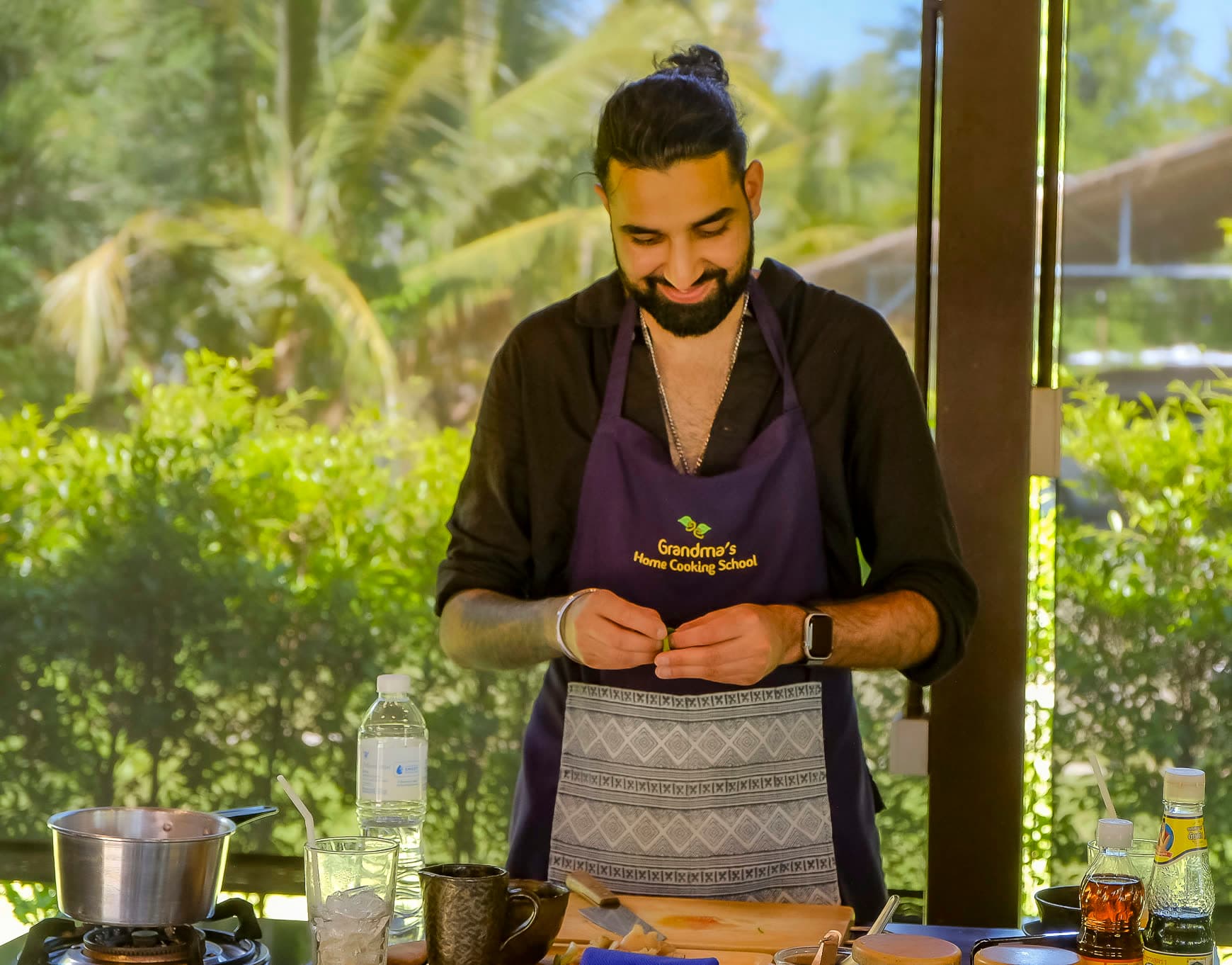 Param in a purple apron smiling down at his hands at a cooking station, palms and hedges behind him
