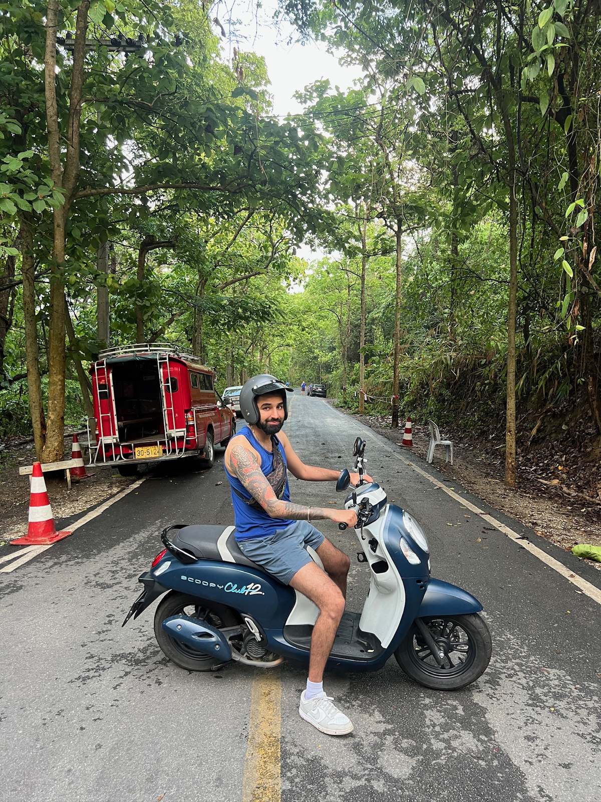 Param in an open-face helmet and a blue vest, astride a blue scooter in the middle of a tree-lined road, a red songthaew parked behind him