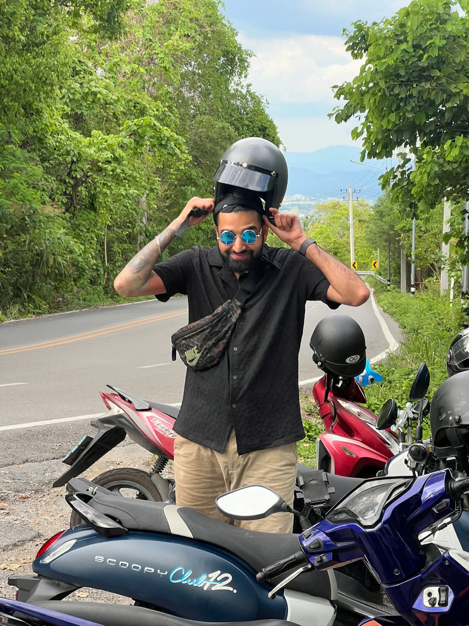 Param in round blue sunglasses pulling off a helmet beside a row of parked scooters on a mountain road