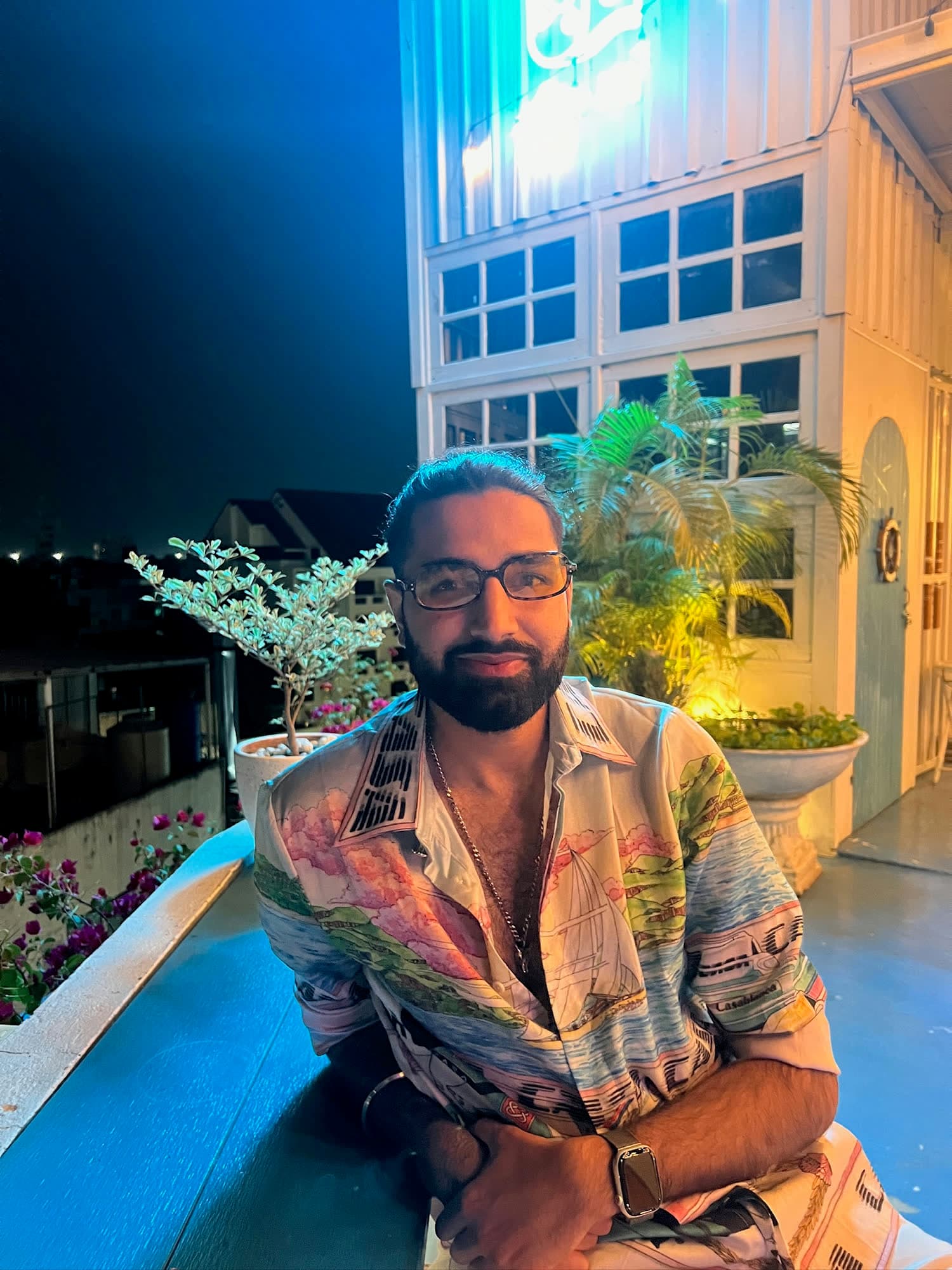 Param in tinted glasses and a printed resort shirt leaning on a ledge on a rooftop terrace at night