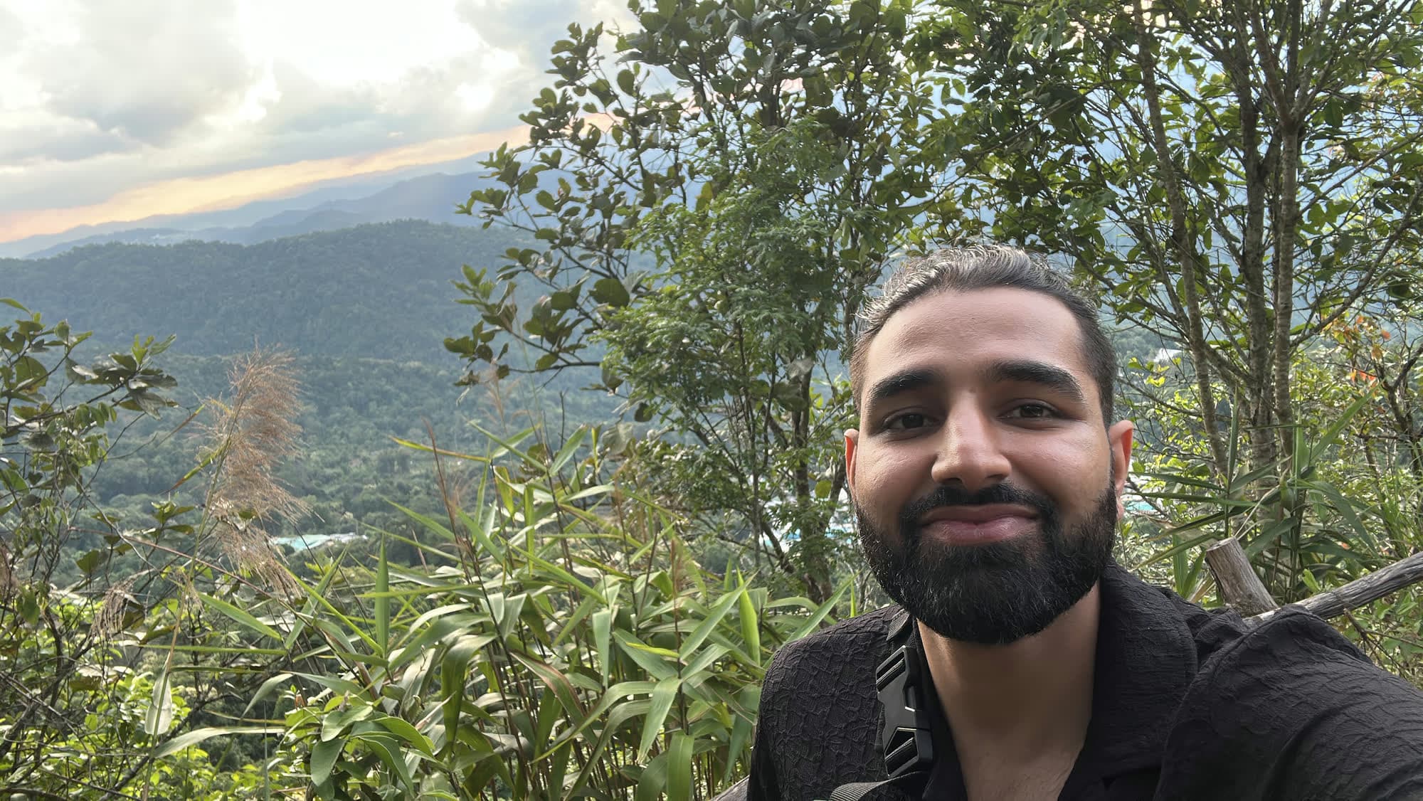 Selfie of Param smiling in front of forested mountain ranges under an evening sky