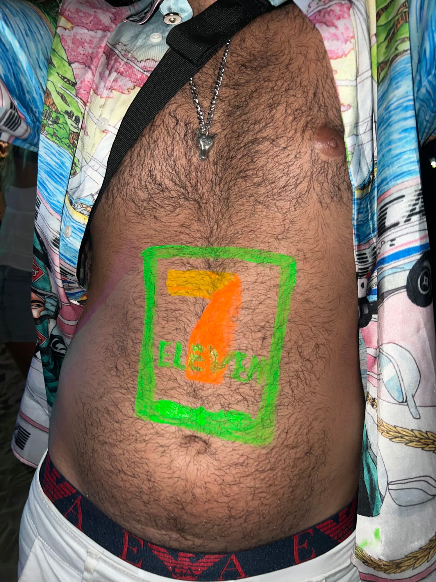 Close-up of a 7-Eleven logo painted in neon green and orange on Param’s stomach, his printed shirt open
