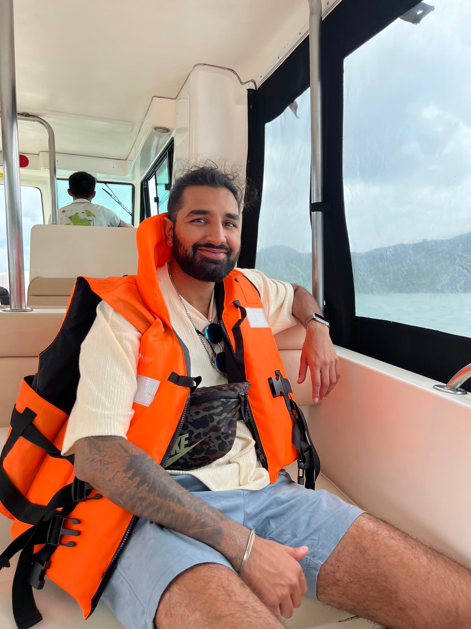 Param in an orange life jacket smiling on the bench seat of a speedboat cabin, sea and hills through the window
