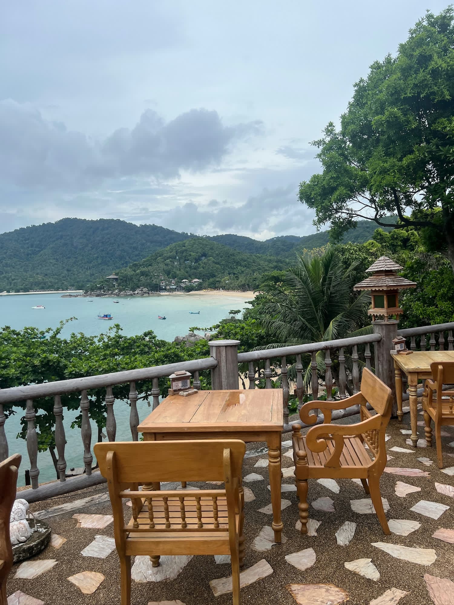 Wooden tables on a stone terrace above a bay with boats and green hills