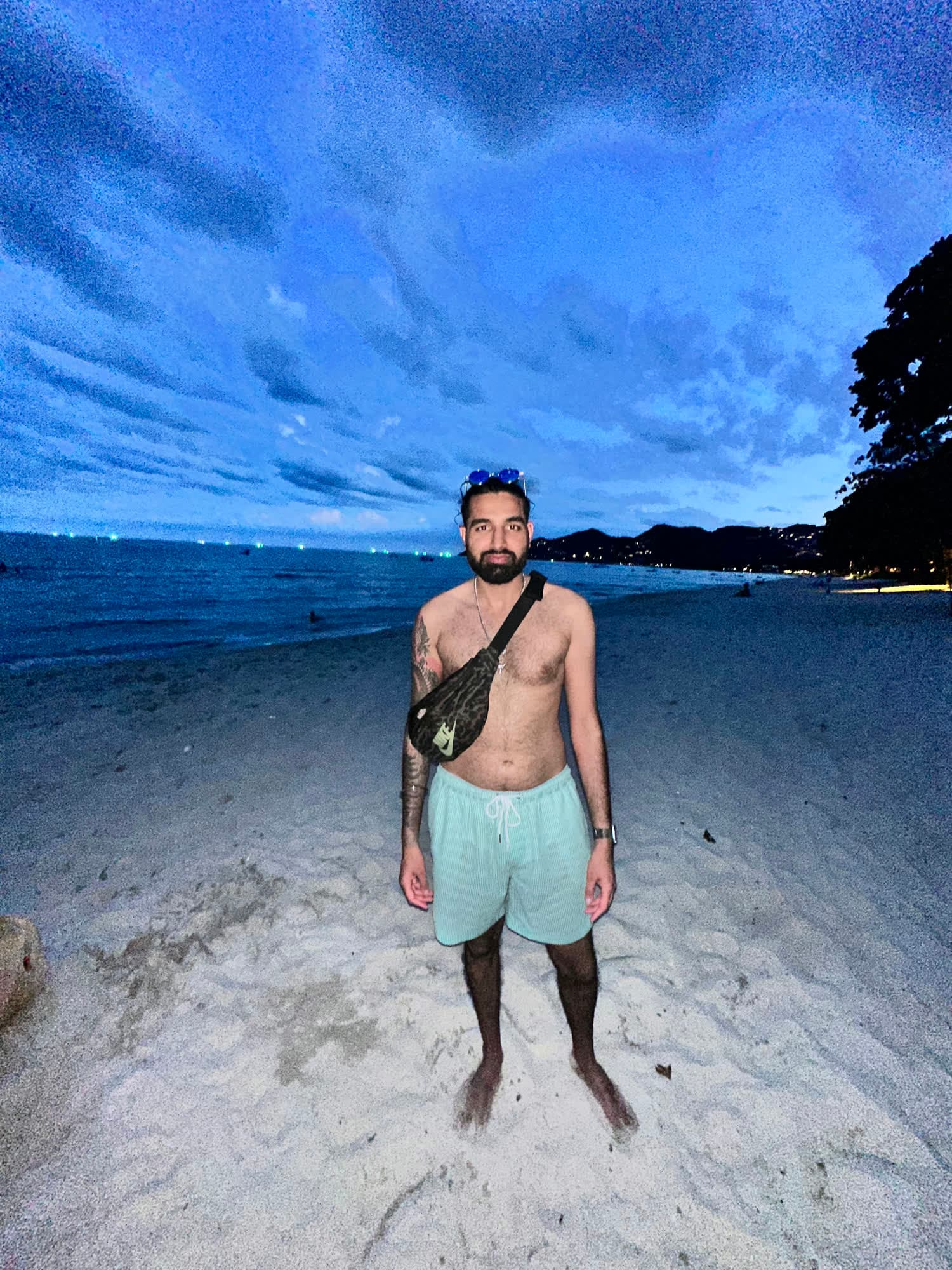 Param shirtless in mint swim shorts standing on a beach at dusk, fishing-boat lights on the horizon