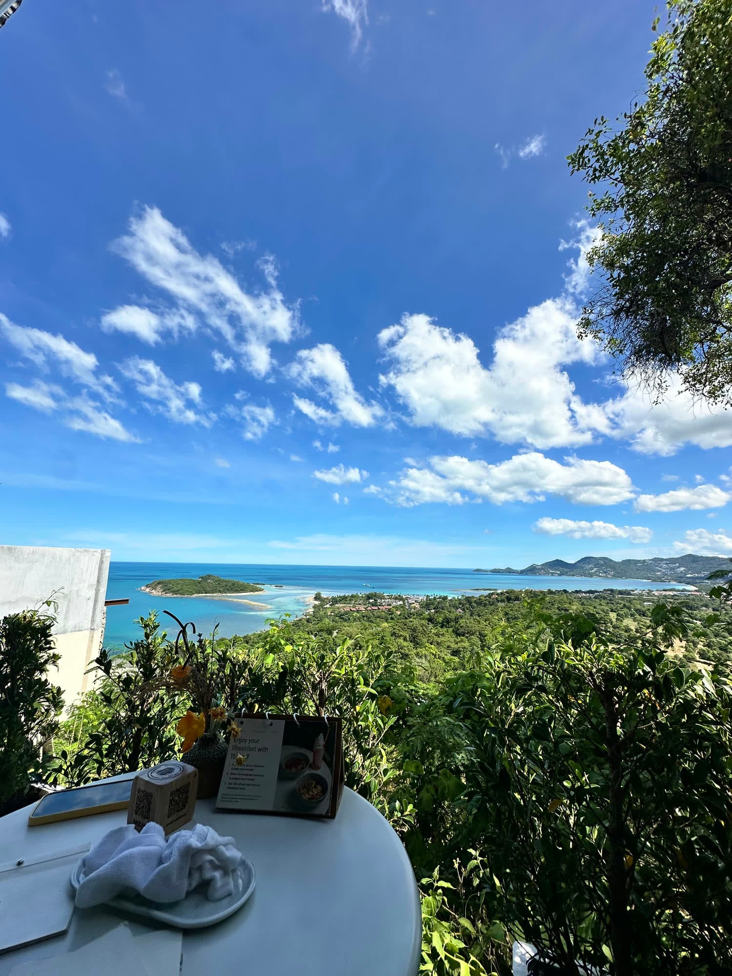 A white table on a hillside terrace looking out over green forest to a turquoise bay and a small island