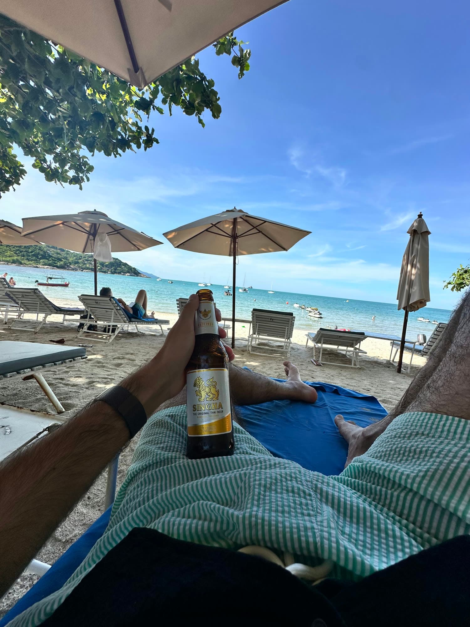 View from a beach lounger: a hand holding a bottle of beer, striped shorts, umbrellas and the sea beyond