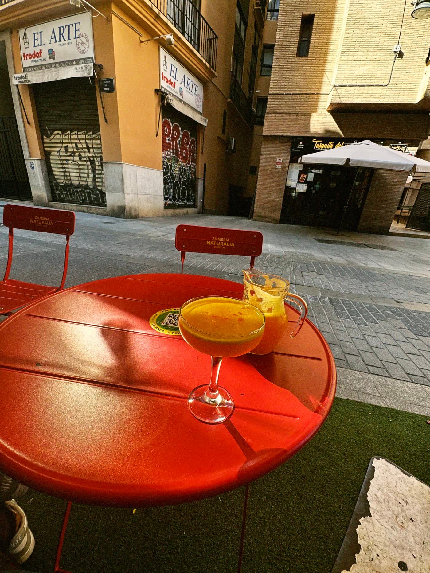Glass of orange juice and a jug on a red café table, a shuttered stamp-maker's shop across the street