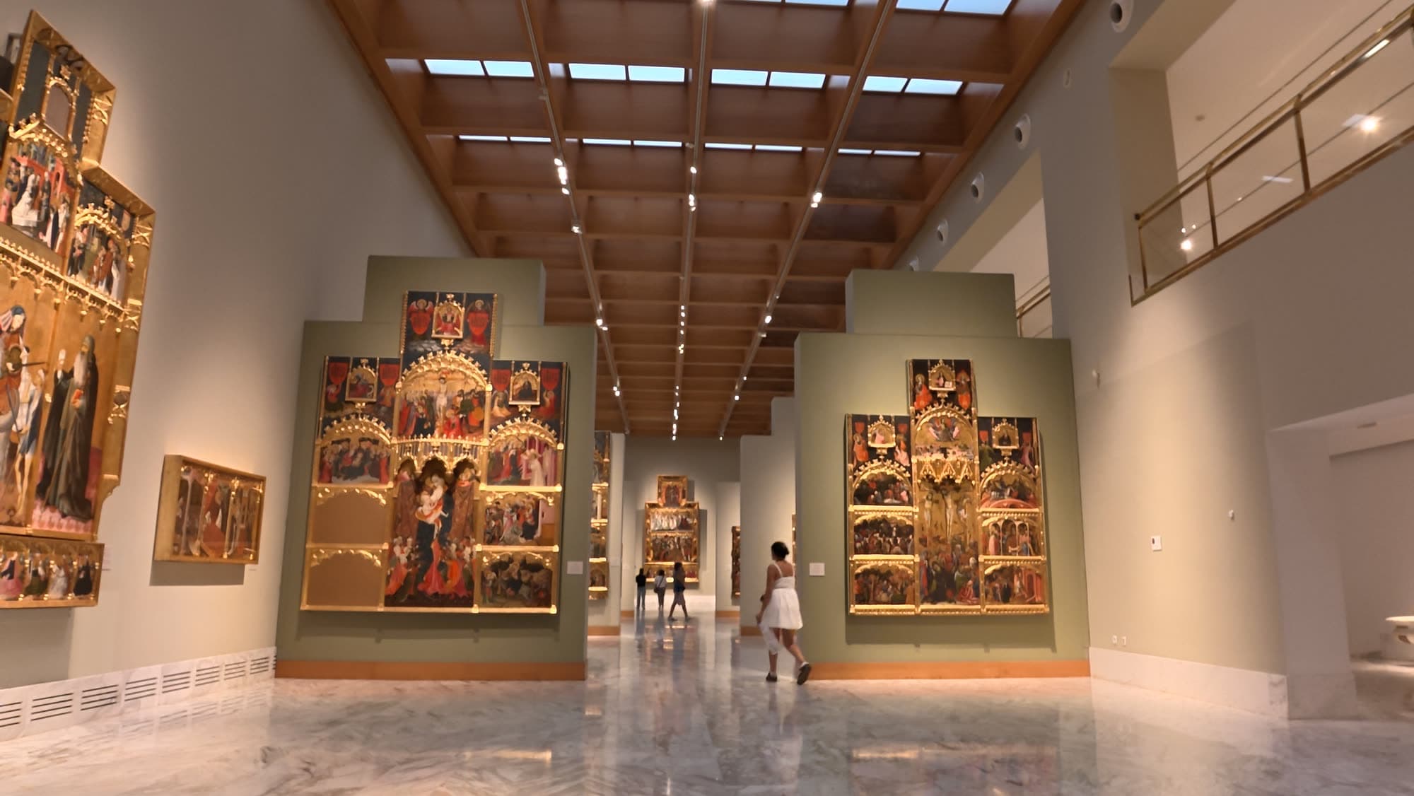 Skylit gallery lined with gilded Gothic altarpieces, one visitor walking away