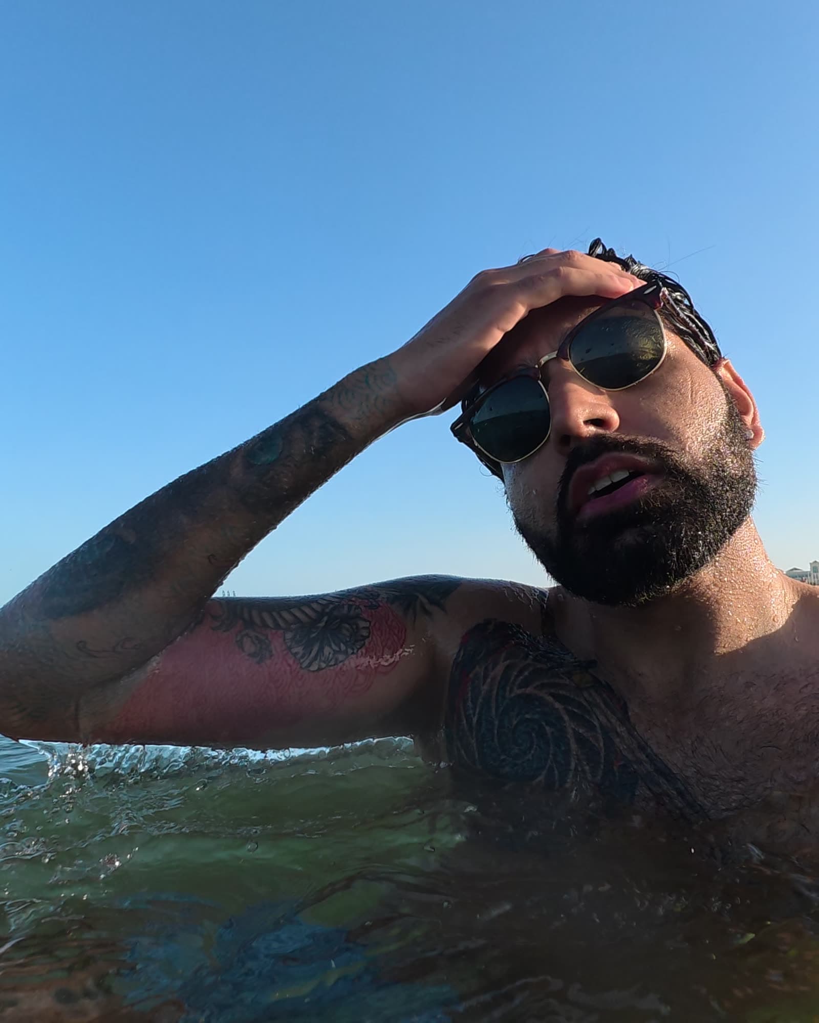 Me shoulder-deep in the sea in sunglasses, one hand on my head, under a clear evening sky