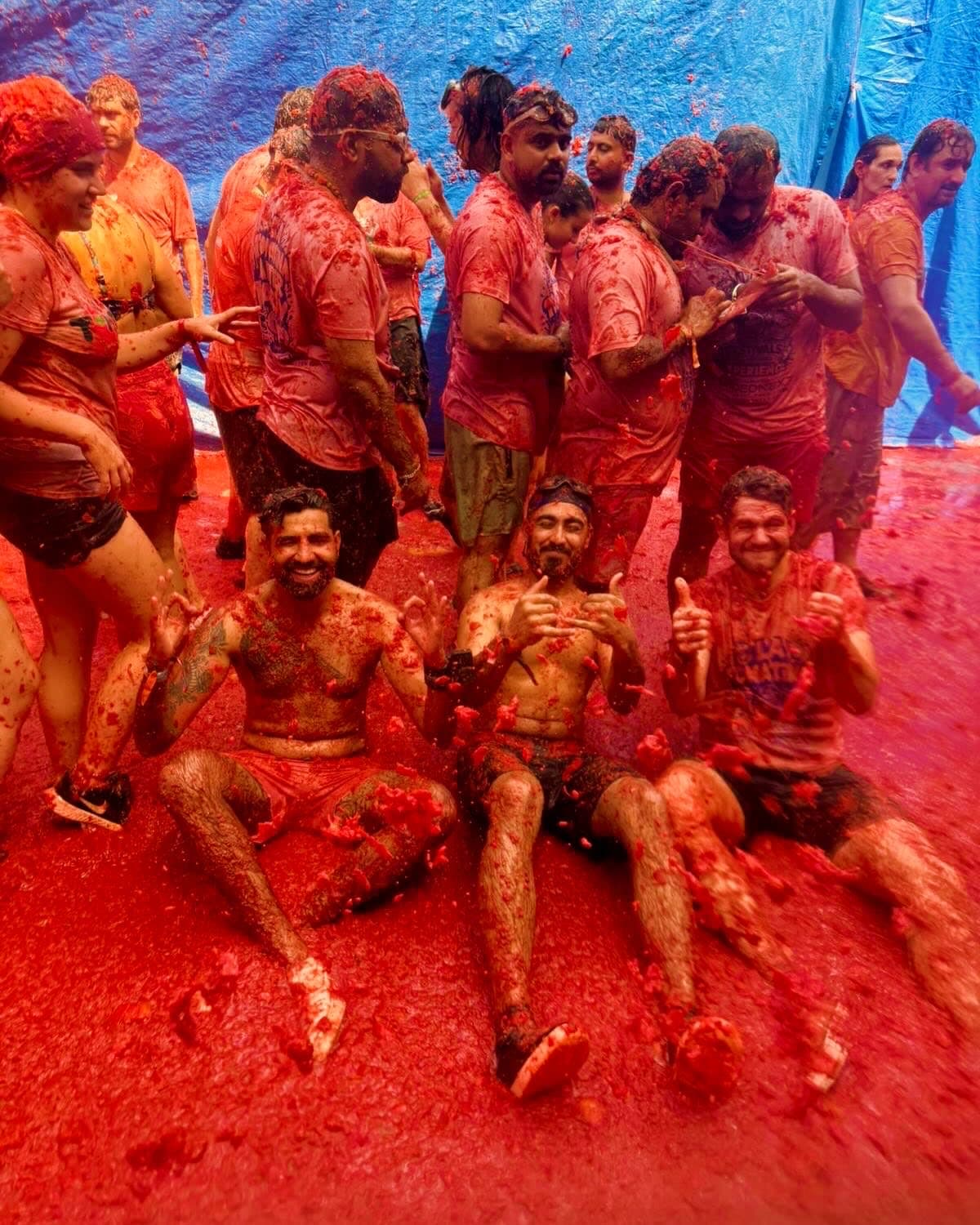 Me and two friends sitting shirtless in the tomato pulp in a Buñol street, grinning and giving the camera hand signs, the crowd standing behind us
