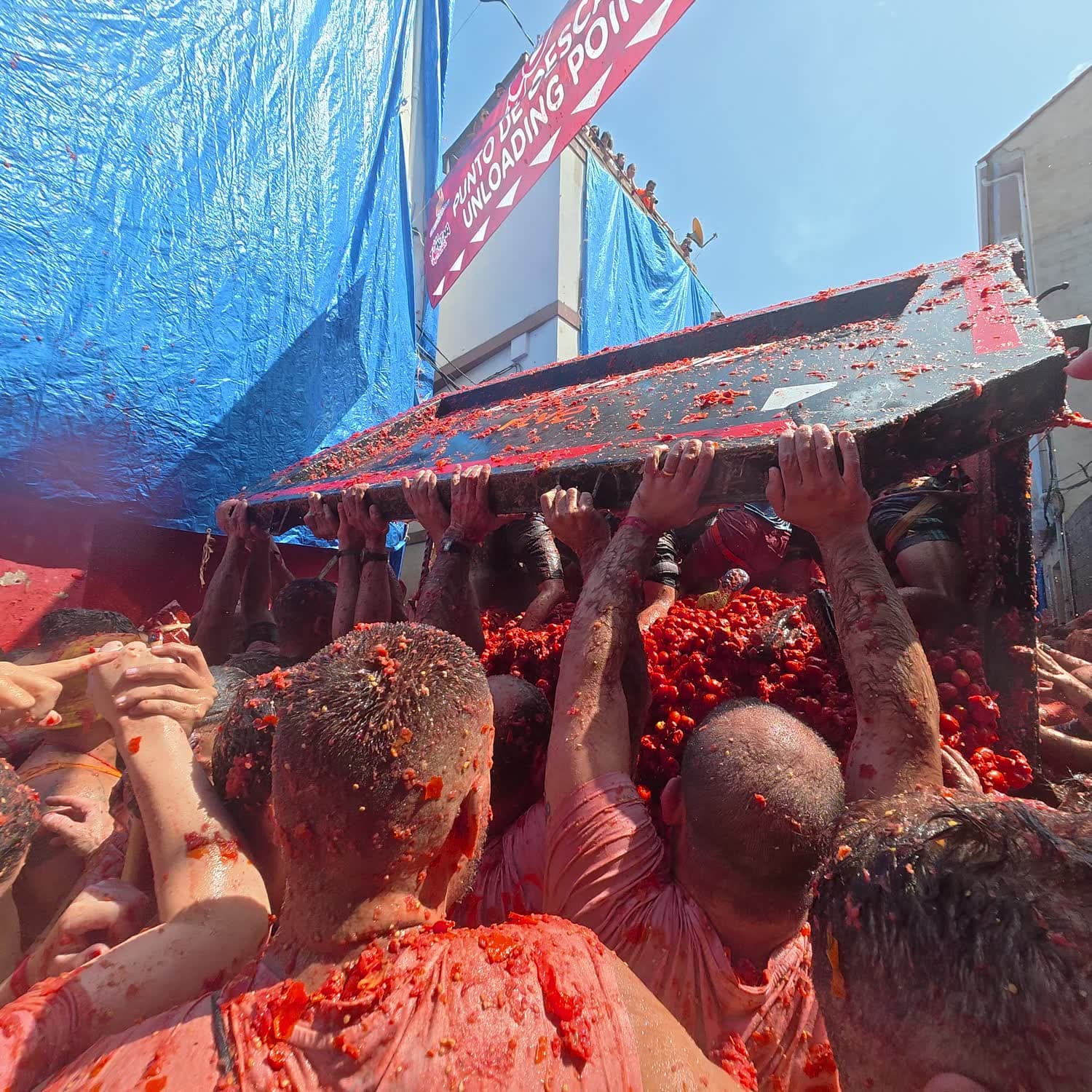 A crowd covered in tomato holding up the tipping bed of a truck as its load spills out